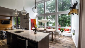 Uma cozinha moderna com uma ilha grande, luminárias pendentes e uma área de jantar com vista para árvores.