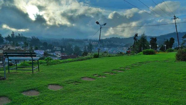 A grassy area with a bench, a paved path, and a distant view of a town and mountains under a cloudy sky. - BRITISH CLIFF CLUB RESORT (Ootacamund)