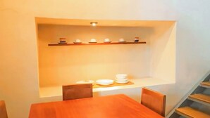 Un coin repas avec une table et des chaises en bois, une étagère intégrée où sont rangés de la vaisselle, et un escalier en arrière-plan.