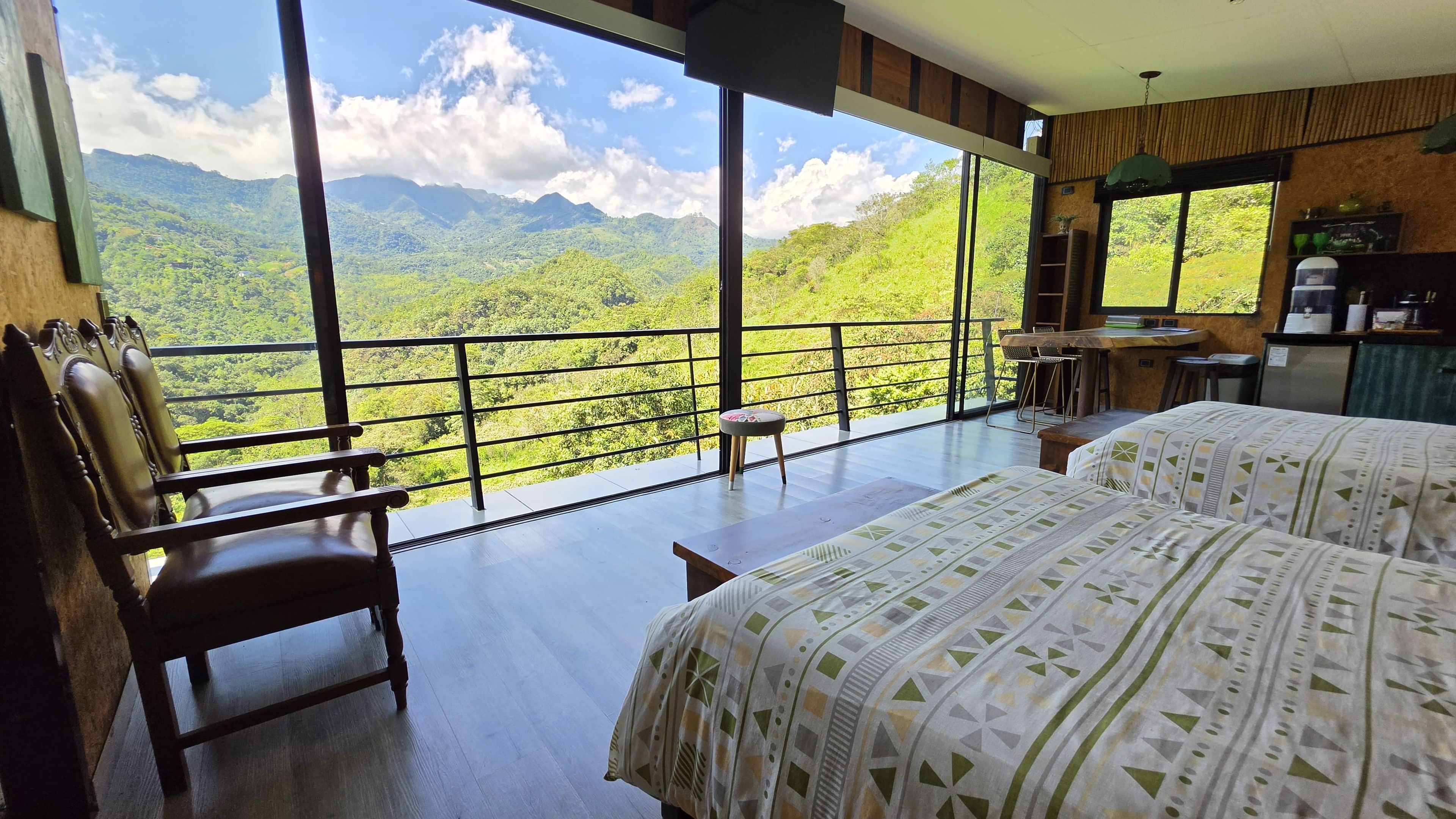 Una habitación con un ventanal que da a un paisaje montañoso, una cama, una silla de madera y una mesita.