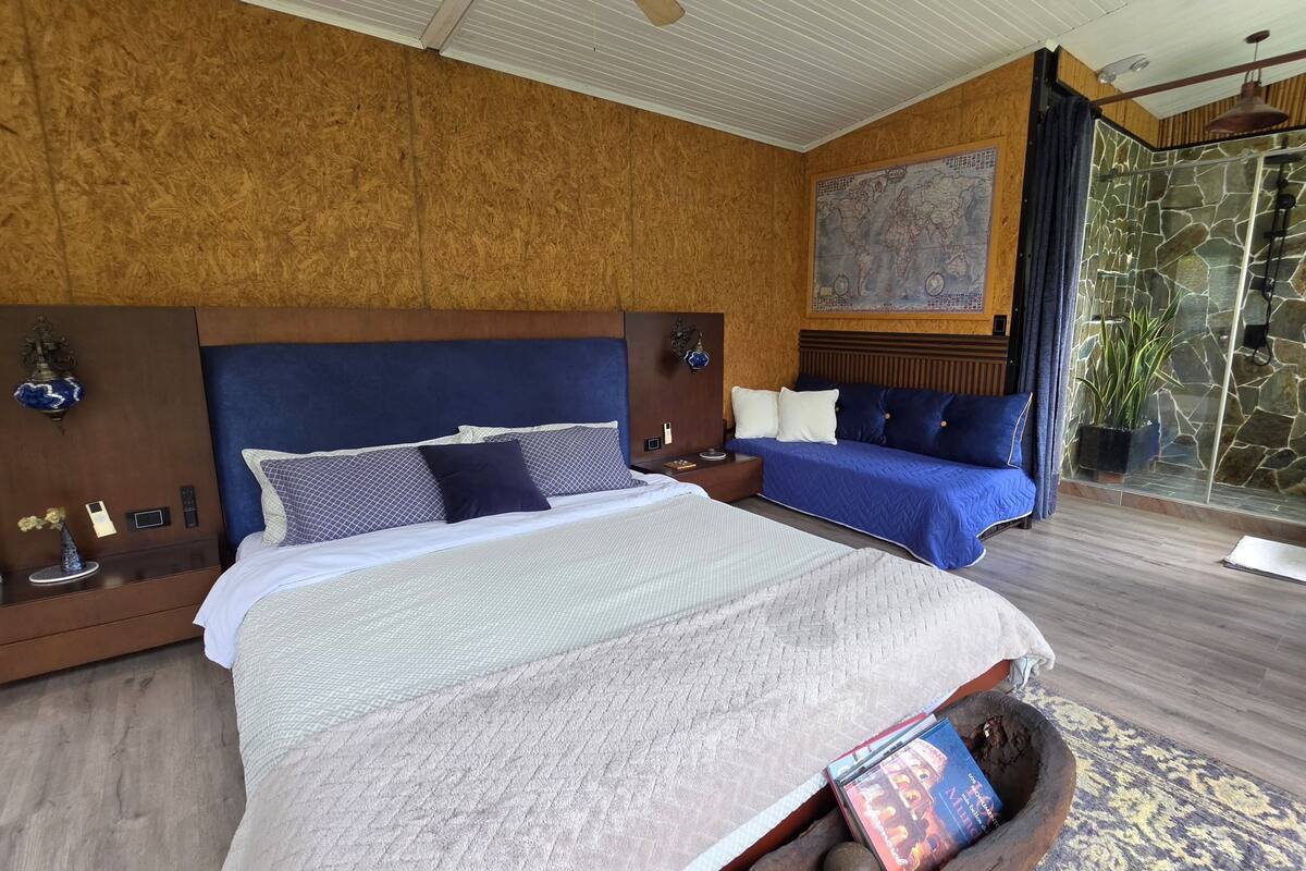 Une chambre avec un grand lit, un petit canapé, une carte du monde accrochée au mur, une douche en pierre et un plafond en bois.