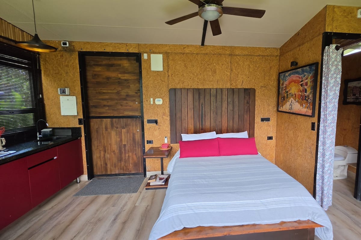 Une chambre d’hôtel de taille réduite, comprenant un lit, un bureau, un ventilateur de plafond et un panneau mural en bois.
