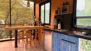 Una cocina con una mesa de madera y taburetes, una ventana con persianas y vistas a árboles.