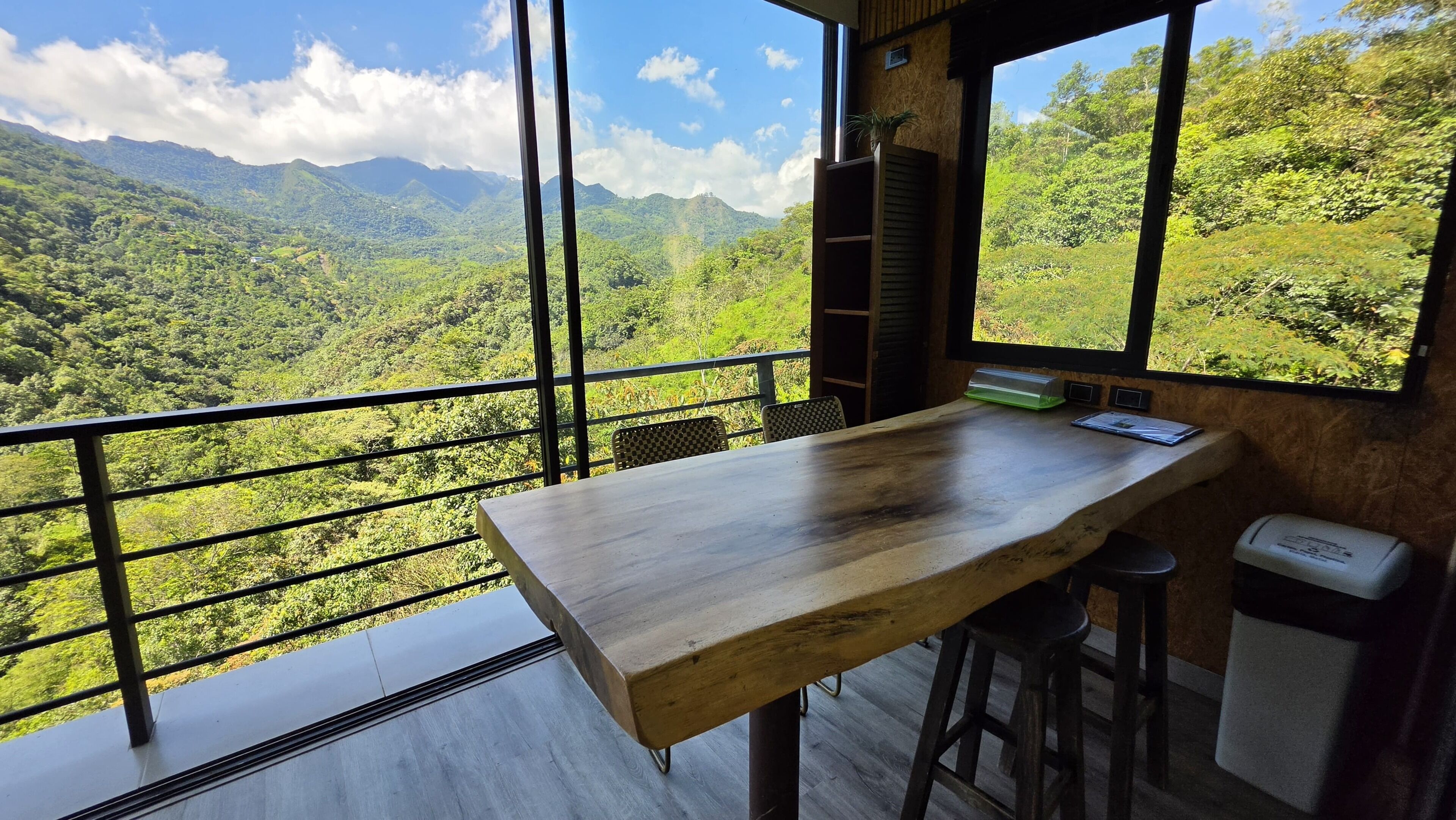 Una habitación con una mesa de madera, taburetes de bar, un ventanal con vistas a una cadena montañosa boscosa y un pequeño cubo de basura.