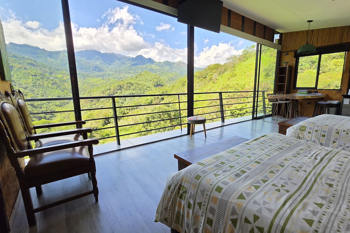 Une pièce dotée d’une grande fenêtre donnant sur un paysage montagneux, avec un lit, une chaise et une petite table.