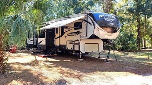 A large RV with a covered living area, a kitchen, and a bathroom, parked in a wooded area with a small table and chairs nearby.
