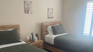 Un dormitorio con dos camas de madera, cada una con cubrecama verde y cabecero de madera. Hay dos mesitas de noche con objetos decorativos y una ventana con persianas.