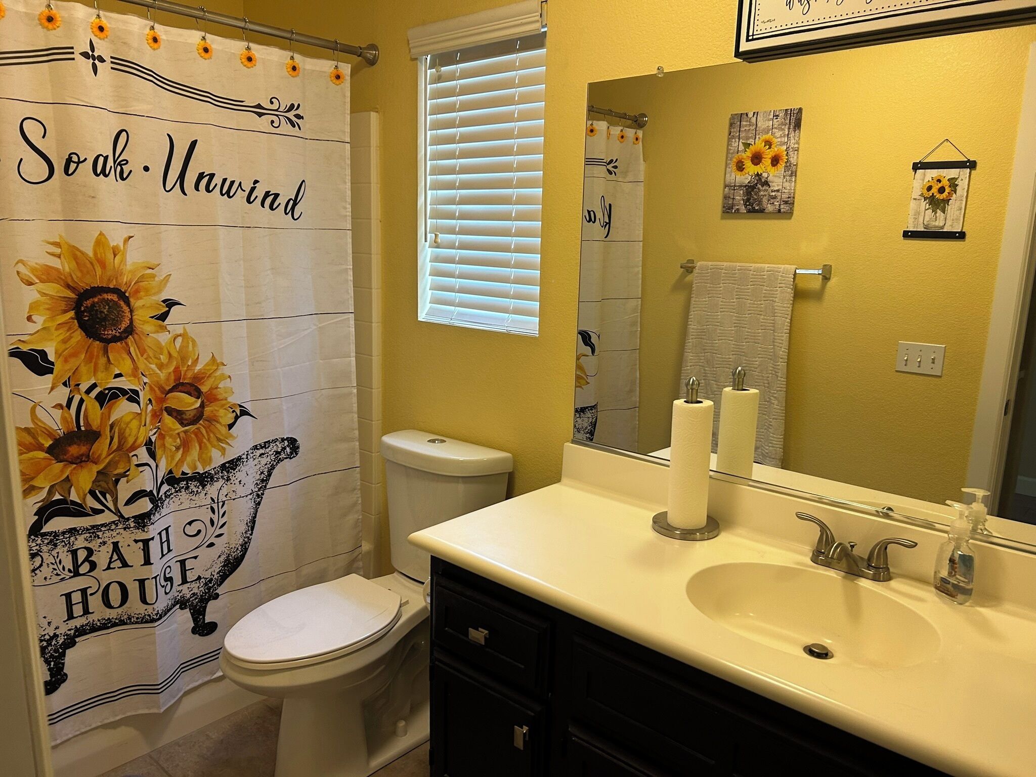 A bathroom with a shower curtain featuring sunflowers and the words "Soak. Unwind" and "Bath House".