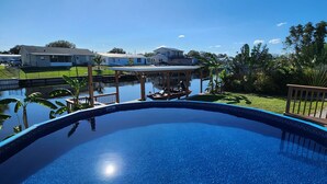 En klarblå pool med en träbrygga och utsikt över hus och grönområden i bakgrunden.