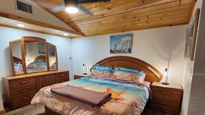 Ett sovrum med ett träpanelat tak, en säng med ett färgglatt överkast, ett sängbord med en lampa, en byrå med en spegel och en tavla på väggen.