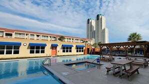 A swimming pool with lounge chairs and a wooden pergola, surrounded by a building with a red roof and blue awnings.