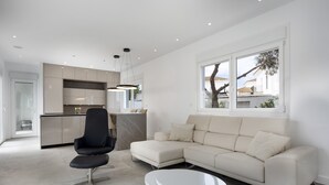 Een moderne woonkamer met een hoekbank, een zwarte fauteuil, een witte salontafel en een keuken op de achtergrond.