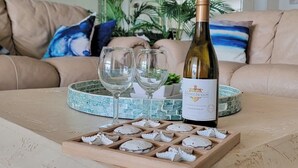 A bottle of wine, two glasses, and a wooden tray with cheese on a table in a living room with a beige sofa and a blue pillow. - High Floor 3BR Corner, Beachfront Saida Towers SPI (South Padre Island)