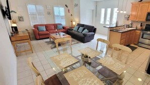 A spacious living area with a sofa, dining table, and chairs, a kitchen with wooden cabinets and appliances, and a large window providing natural light.