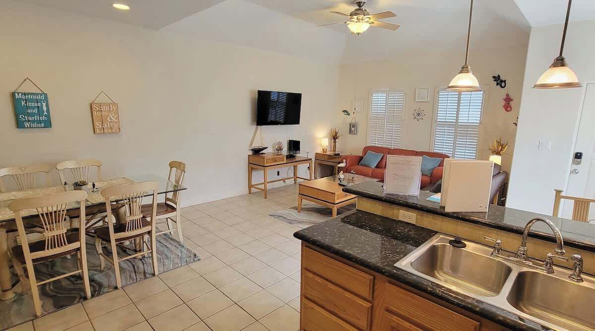 A spacious living area with a dining table, chairs, a sofa, and a TV. There is a kitchen with a sink and cabinets.