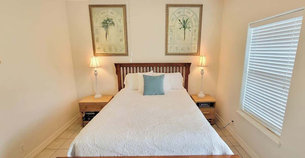 A neatly made bed with a white bedspread and a single blue pillow, flanked by two bedside tables with lamps, in a room with a large window and two framed botanical prints on the wall.