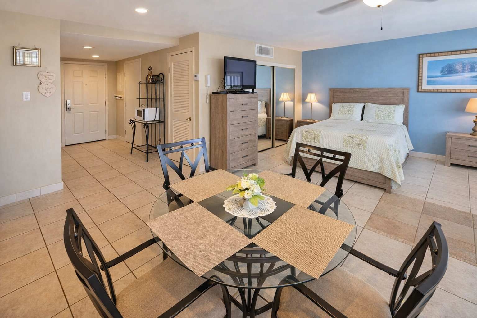 A hotel room with a bed, a TV, a dining area with chairs and a table, and a kitchenette.