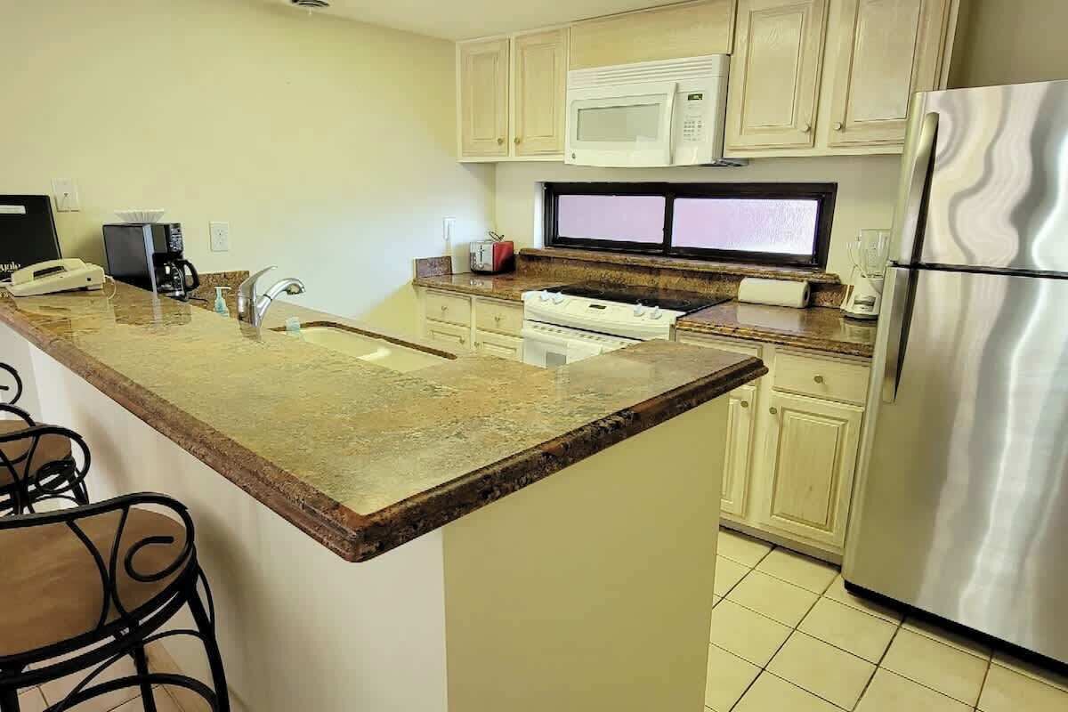 Una cocina con encimera de granito, armarios blancos, refrigerador de acero inoxidable, microondas, estufa y pileta.