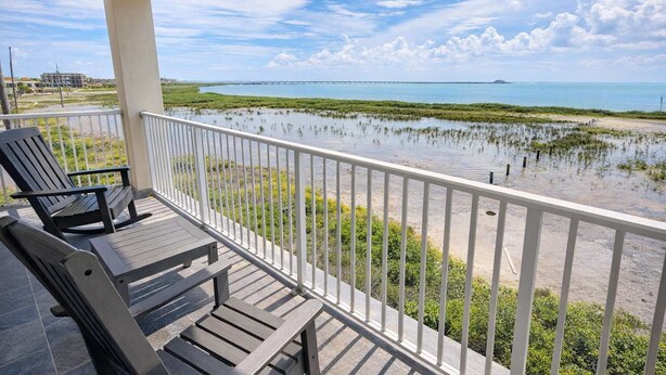 A balcony with two rocking chairs overlooking a coastal view with a bridge in the distance. - 3-Level Bayfront | Boat Dock | Walk-In Pool | SPI (South Padre Island)