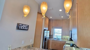 A modern kitchen with wooden cabinets, a central island, and pendant lighting. A sign on the wall reads "MY HEART IS IN TEXAS". - 3-Level Bayfront | Boat Dock | Walk-In Pool | SPI (South Padre Island)