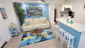 Una sala de estar moderna con sofá, mesa de centro y taburetes de bar. Se observa una cocina al fondo.