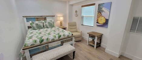 A bedroom with a bed, a chair, a small table, and a window with blinds.