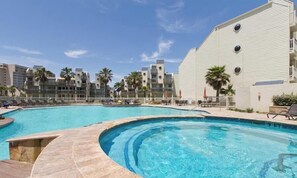 Uma piscina com design curvo, cercada por um edifício branco com varandas e palmeiras.