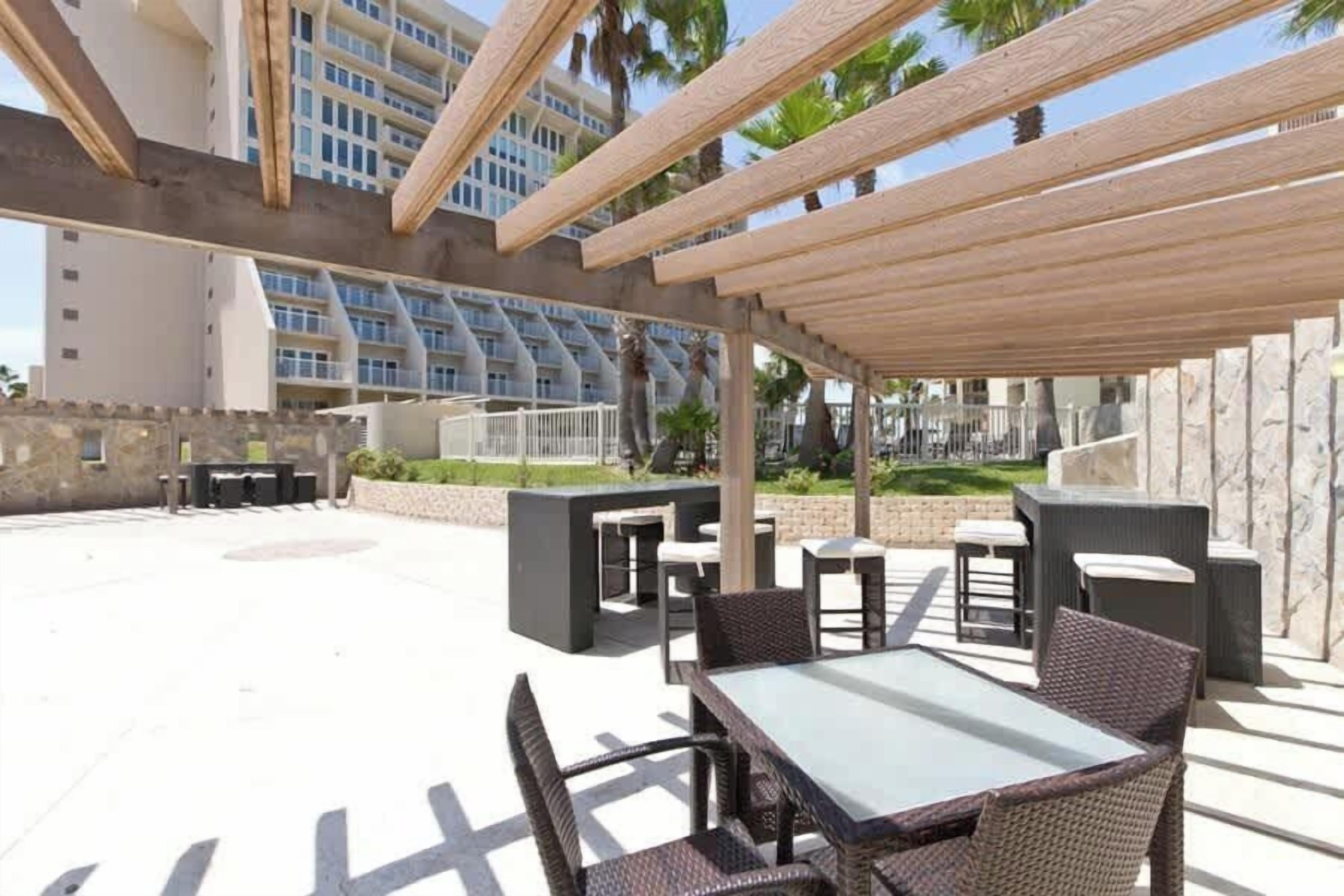 Un espacio patio al aire libre con mesas y sillas, una estructura de pérgola y vistas a un edificio en el fondo.