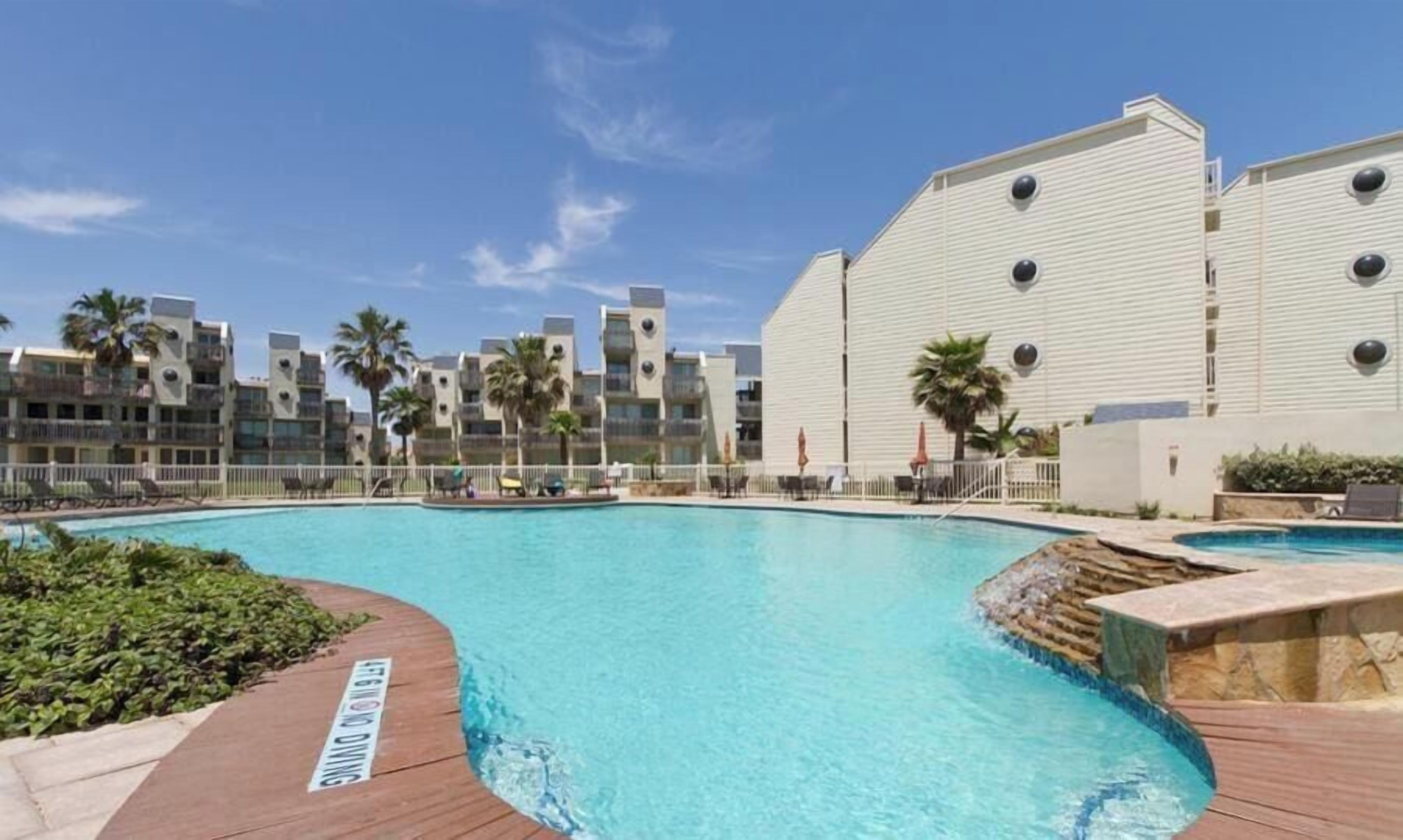 Una piscina con plataforma de madera, rodeada de edificios de varios pisos con balcones y palmeras.