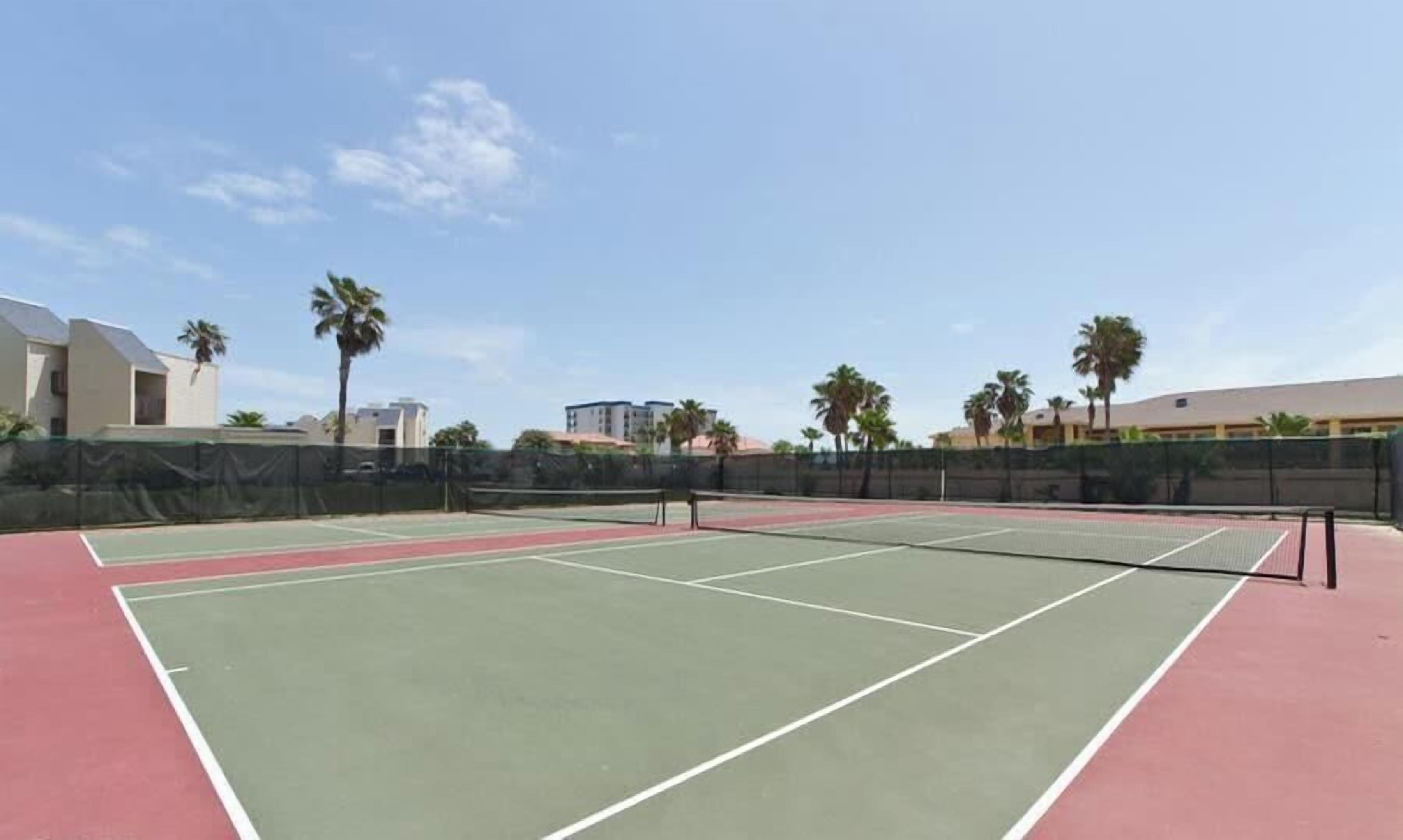 Ein Tennisplatz im Freien mit grüner und roter Belag, umgeben von einem Zaun und Palmen.