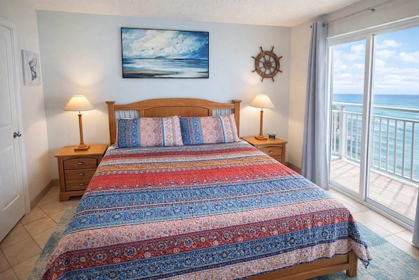 A bedroom with a bed, two bedside tables, a nightstand, a window with a view of the ocean, a painting on the wall, and a ship's wheel decoration.
