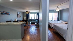 Una habitación de hotel compacta con kitchenette, sofá, cama y ventilador de techo.