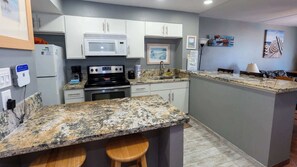 A kitchen with white cabinets, a granite countertop, a stove, and a microwave. There are two stools at the counter.