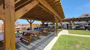 An outdoor covered patio with picnic tables and chairs, a wooden fence, and a grassy area.