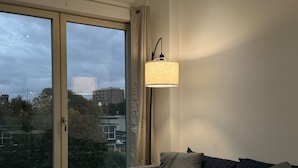 A bedroom with a large bed, a grey sofa, a floor lamp, and a sliding glass door leading to a balcony.