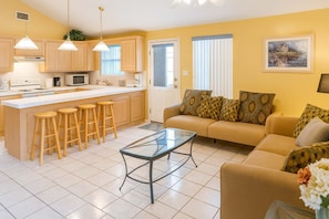 A modern kitchen with a central island, bar stools, a glass coffee table, and a living area with a sofa and patterned cushions.