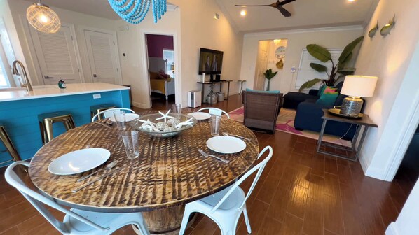 A dining area with a round table set for four, a kitchen in the background, and a living area with a sofa and plants.