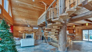 L’intérieur d’une cabane en rondins, avec un escalier en colimaçon, une cheminée, un coin repas et une grande fenêtre donnant sur un paysage enneigé.