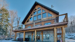 Une cabane en rondins à deux étages, dotée d’un balcon, de fondations en pierre et d’un télésiège visible au loin.