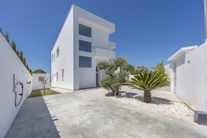 Un edificio moderno blanco con techo plano, un camino de hormigón y pequeños árboles.