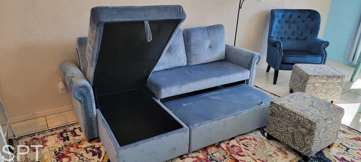 Una habitación con un sofá que se convierte en cama, un reposapiés estampado y un sillón azul.