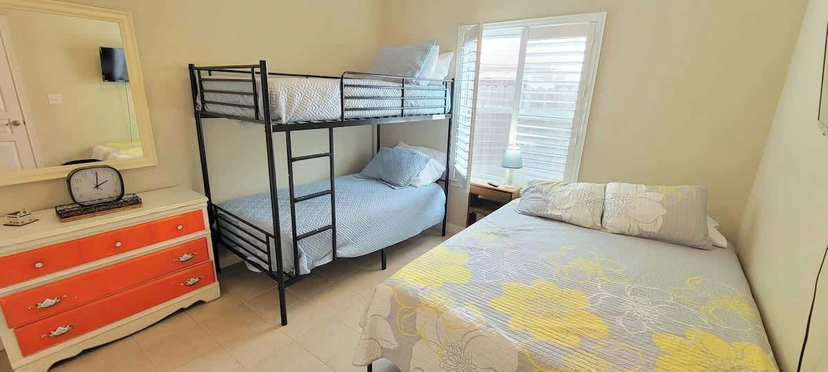 Une chambre avec un lit superposé, un lit simple, une commode avec des tiroirs, une table de chevet avec une lampe et une fenêtre avec des stores.