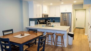 A modern kitchen with white cabinets, a stainless steel refrigerator, a microwave, and a coffee maker. There is a wooden dining table with four chairs and a small island with stools. - Bahia Mar Beachfront "Remodel " Water Views (South Padre Island)