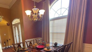 A dining room with a wooden table set for six, surrounded by chairs. A chandelier hangs above the table, and there is a large window with blinds to the right.