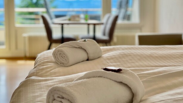 A neatly made bed with rolled towels and a view of a dining area through a large window. - Modern "Waldluft" apartment with pool and sauna in Scheidegg in the Allgäu (Scheidegg)