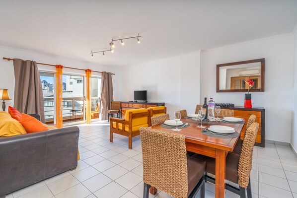 Ein modernes Wohnzimmer mit Sofa, Esstisch, Stühlen, Fernseher und einem Balkon mit Vorhängen.