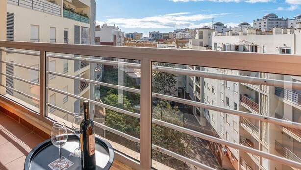 A balcony with a glass table, a bottle, and a glass, overlooking a cityscape with buildings and a clear sky. - Apartment 'Cool Motion Rocha Beach T1' with Balcony, Wi-Fi and Air Conditioning (Portimão)