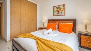 A neatly made bed with a white and yellow blanket, orange pillows, and a wooden headboard. There is a painting on the wall above the bed and a wooden wardrobe to the left. - Apartment 'Cool Motion Rocha Beach T1' with Balcony, Wi-Fi and Air Conditioning (Portimão)