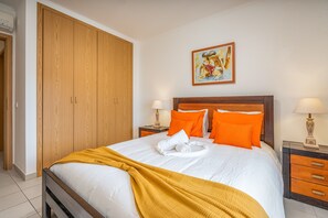 Una cama bien hecha con manta blanca y amarilla, almohadas naranjas y cabecera de madera. Hay un cuadro en la pared sobre la cama y un armario de madera a la izquierda.