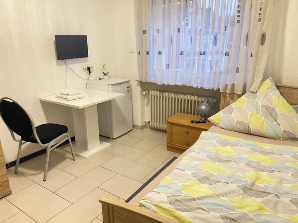 A bedroom with a bed, a desk with a chair, a radiator, and a window with curtains. - Triebenerhof Hotel (Quierschied)
